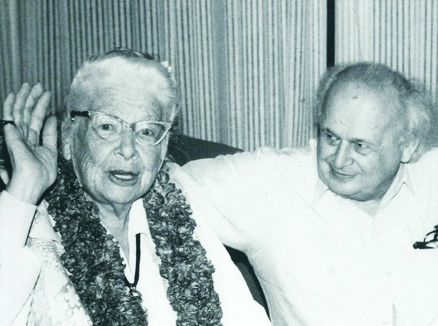 Ida Rolf, white-haired, in cat-eye glasses and a thick garland, raises one hand as she speaks; Moshe Feldenkrais sits beside her with his arm behind her and smiles, before a striped curtain