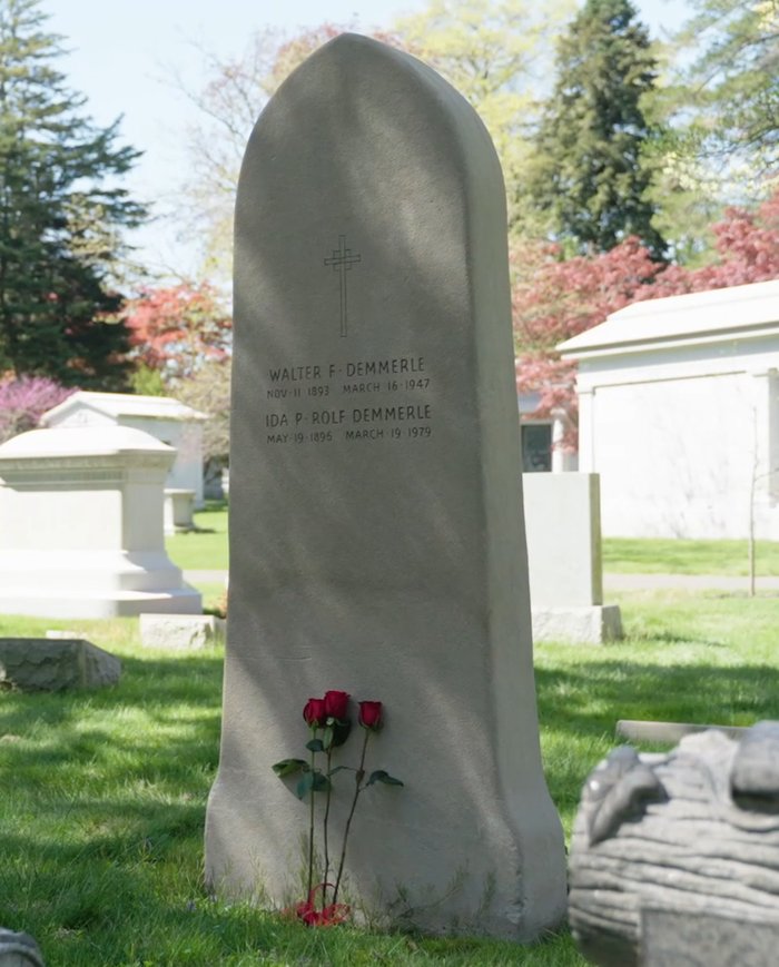 A tall pale headstone with a rounded peak, carved with a cross and the names of Walter F. Demmerle and Ida P. Rolf Demmerle, three red roses at its foot, on a spring lawn among mausoleums
